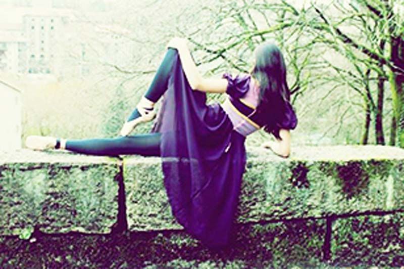 Una persona con un vestido morado está recostada sobre un muro de piedra al aire libre, entre árboles sin hojas.