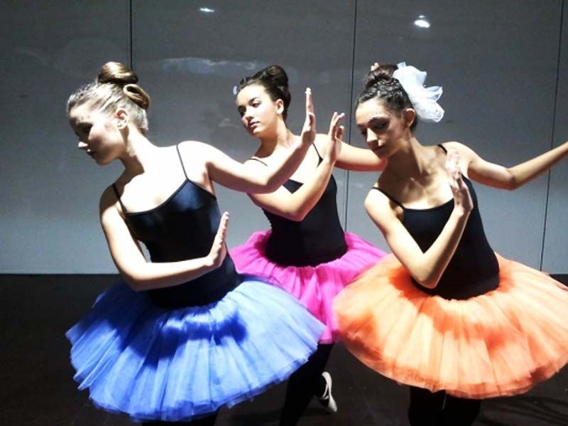 Tres bailarinas de ballet posando con tops negros y tutús azules, rosas y naranjas en un escenario oscuro.