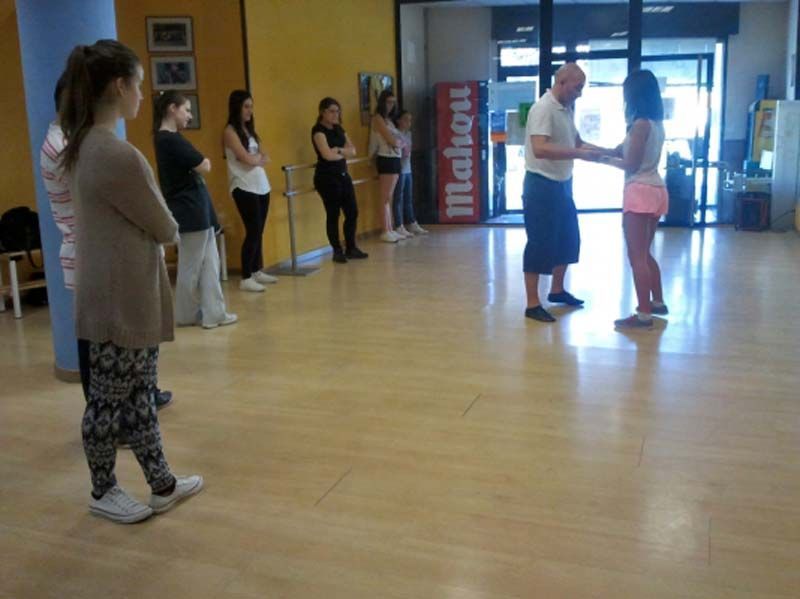 Varias personas se encuentran en un estudio de baile, mientras dos de ellas practican una postura en pareja cerca de la entrada.