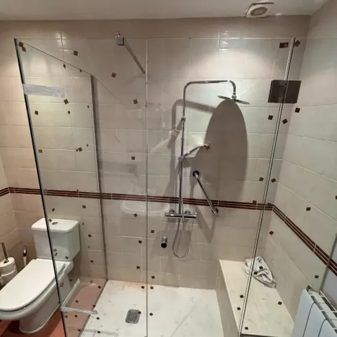 Baño accesible con inodoro, ducha a ras de suelo y puertas de ducha de cristal plegables.