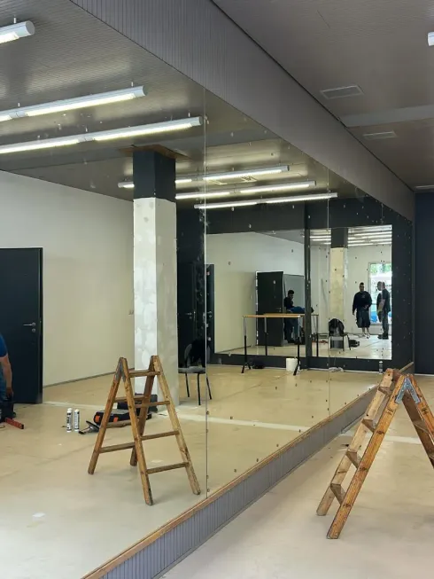 Estudio de danza con espejos, suelo de madera, escaleras y luces fluorescentes; algunas personas están de pie al fondo.