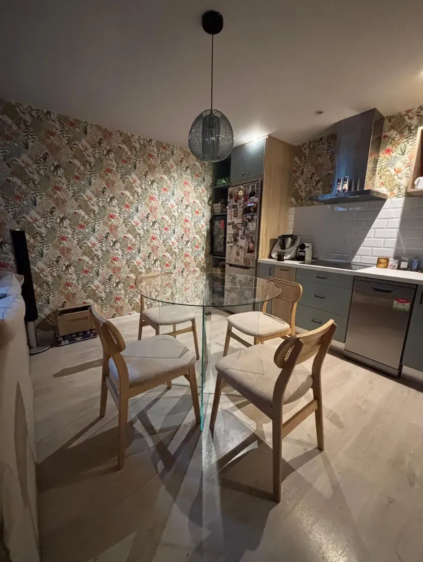 Pequeño comedor con papel pintado floral, sillas de madera y una cocina a lo largo de la pared derecha.