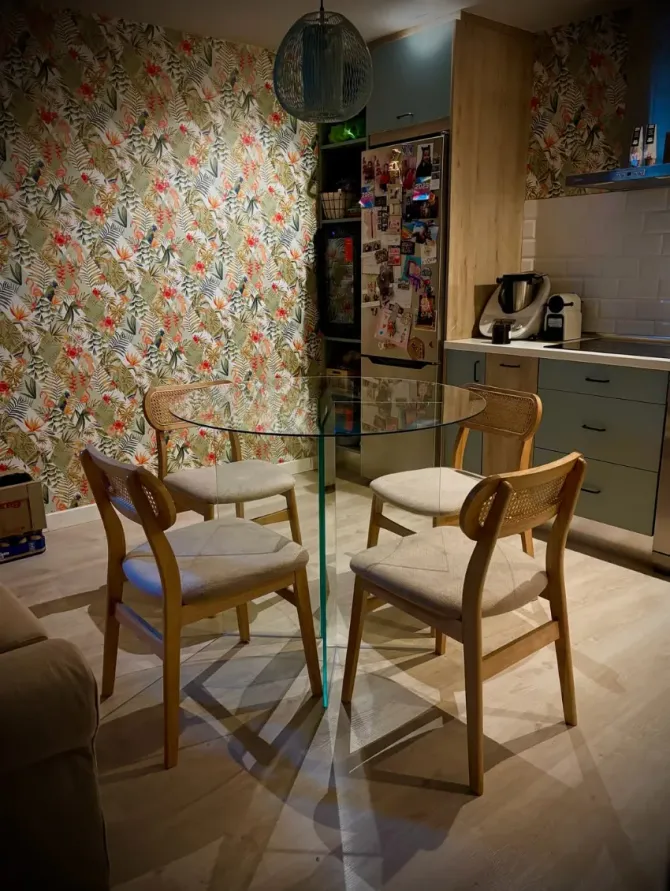 Pequeño comedor con papel pintado floral, mesa redonda de cristal y cuatro sillas de madera cerca de una encimera de cocina.