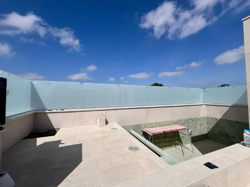 Terraza en la azotea con suelo de piedra clara, barandillas de cristal y una pequeña zona de estar bajo un cielo azul.