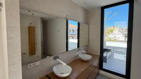 Tocador de baño moderno con dos lavabos, espejos grandes y una puerta de cristal que da a una terraza soleada.