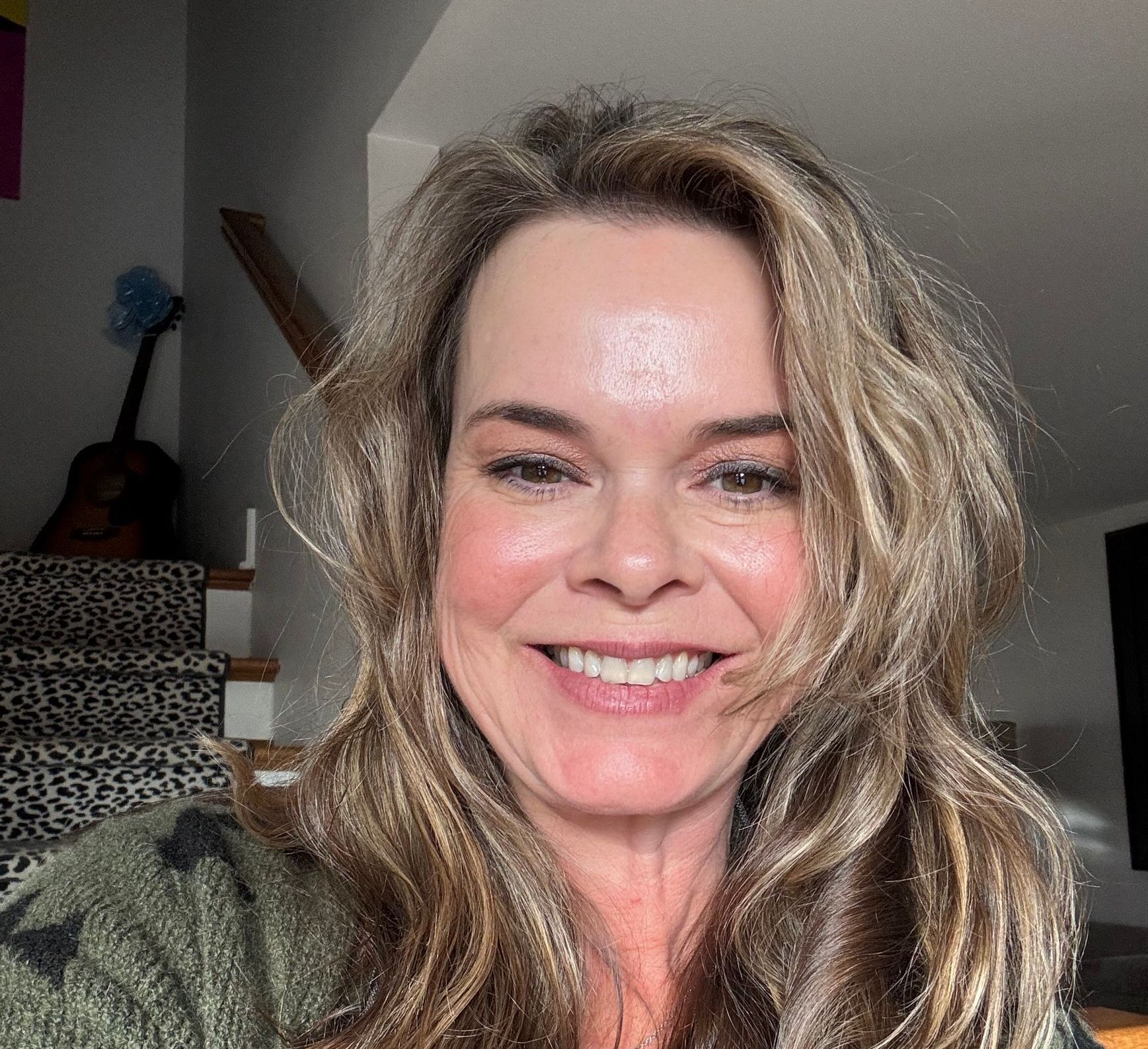 Woman smiling, light brown hair, outdoors, wearing a green patterned top, bright day.