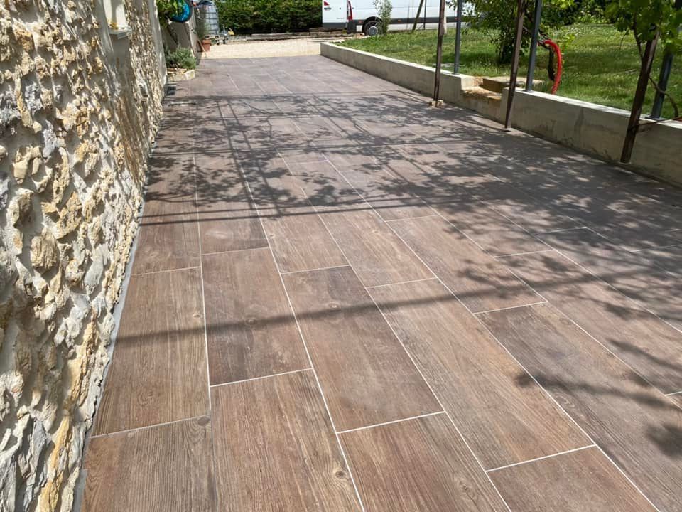 Allée en pierre longeant un mur et une pelouse, avec des arbres à l'ombre et un chemin pavé menant au loin.