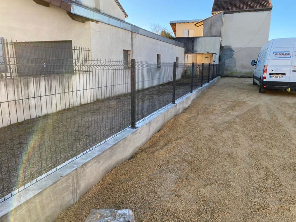 Une ruelle étroite en gravier longe des bâtiments blancs et une clôture en fil de fer, avec une camionnette blanche garée au bout.