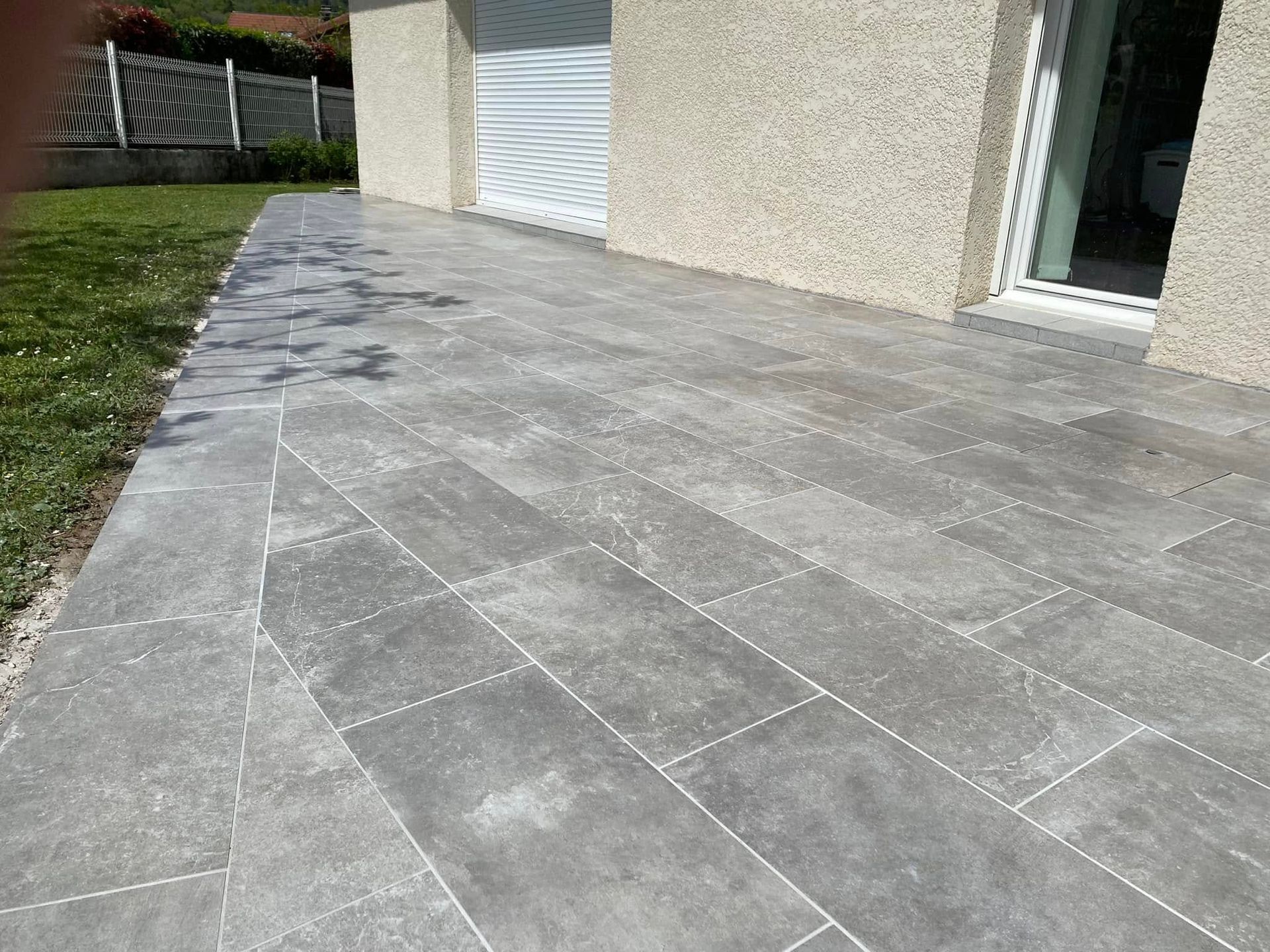 Terrasse en pierre grise bordant une pelouse et une maison, avec une porte de garage blanche en arrière-plan.