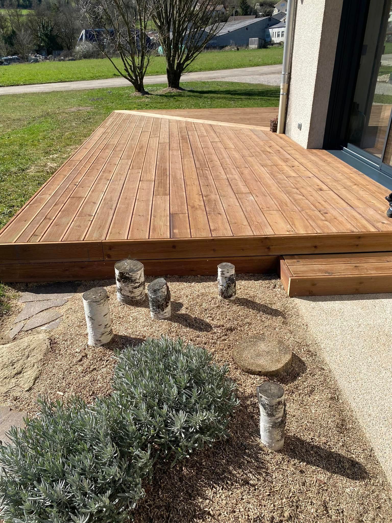 Terrasse en bois surplombant une cour à côté d'une maison, avec un petit arbuste et des bornes de chemin en béton.