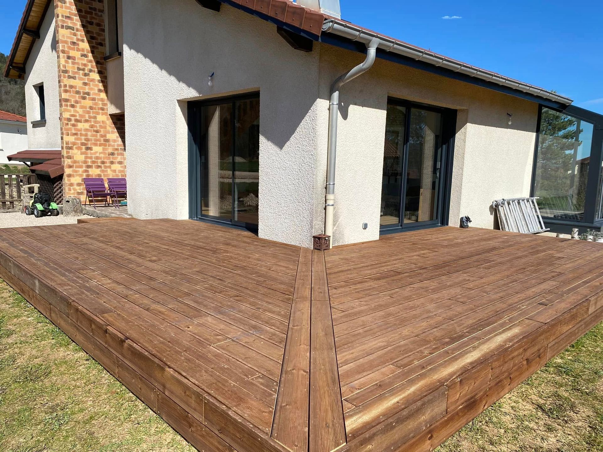 Une terrasse en bois entoure une maison blanche avec de grandes portes vitrées et une cheminée en briques.