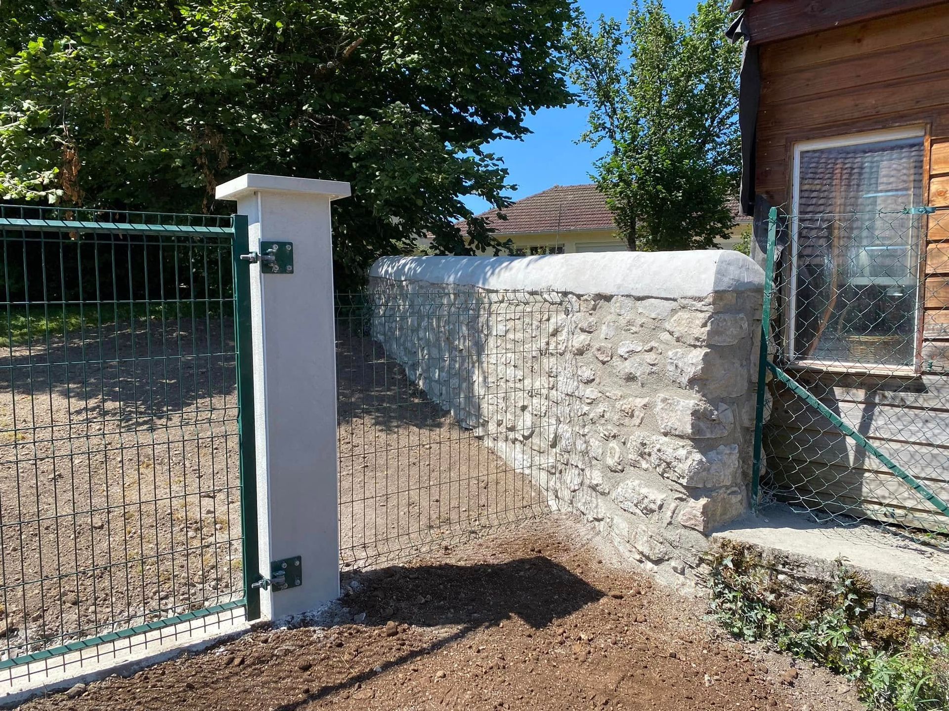 Portail en pierre et pilier blanc bordant une allée de gravier et un bâtiment en bois dans une cour ensoleillée