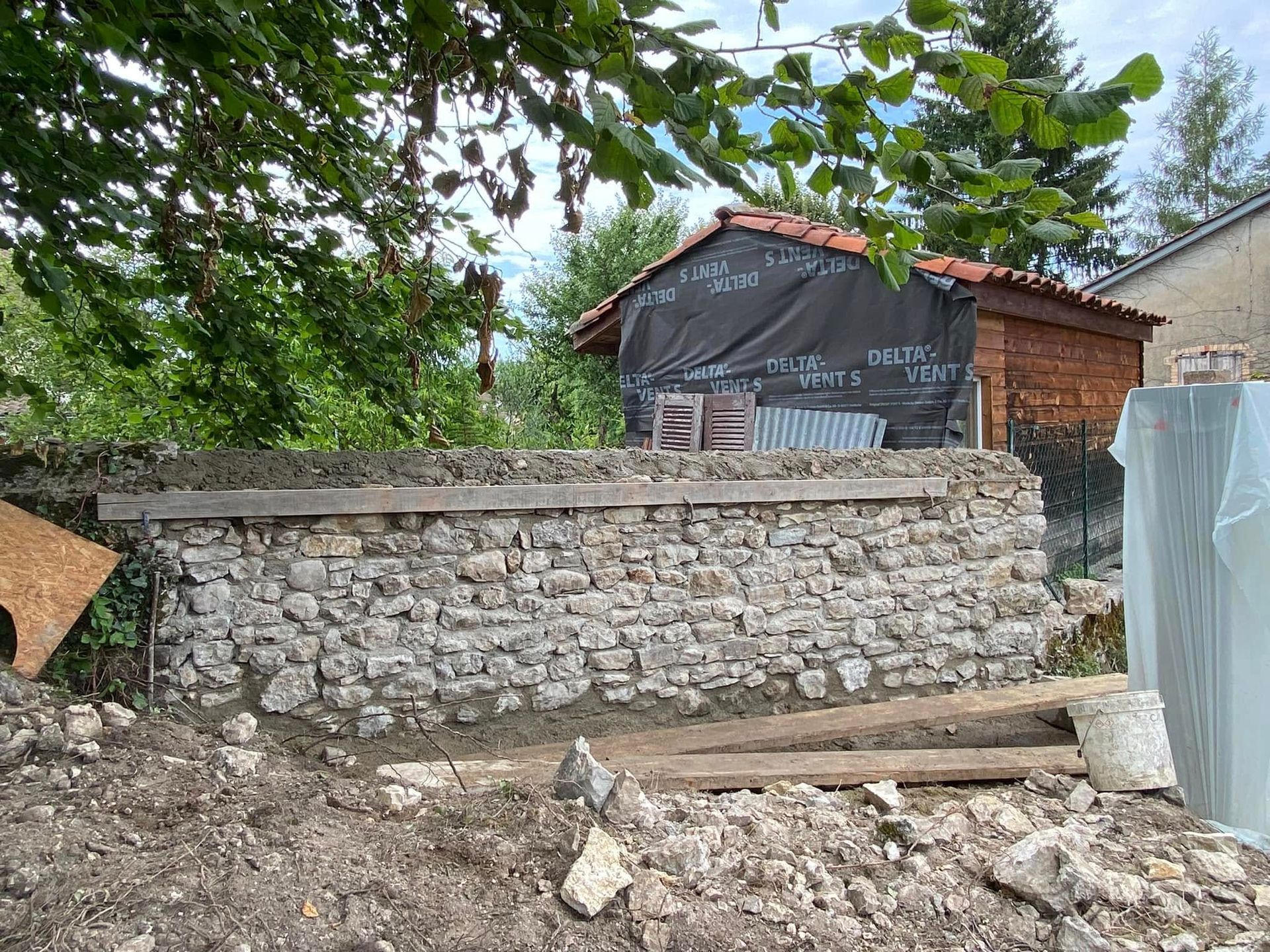 Mur de soutènement en pierre bordant un chemin de terre, avec une petite maison et des arbres derrière.