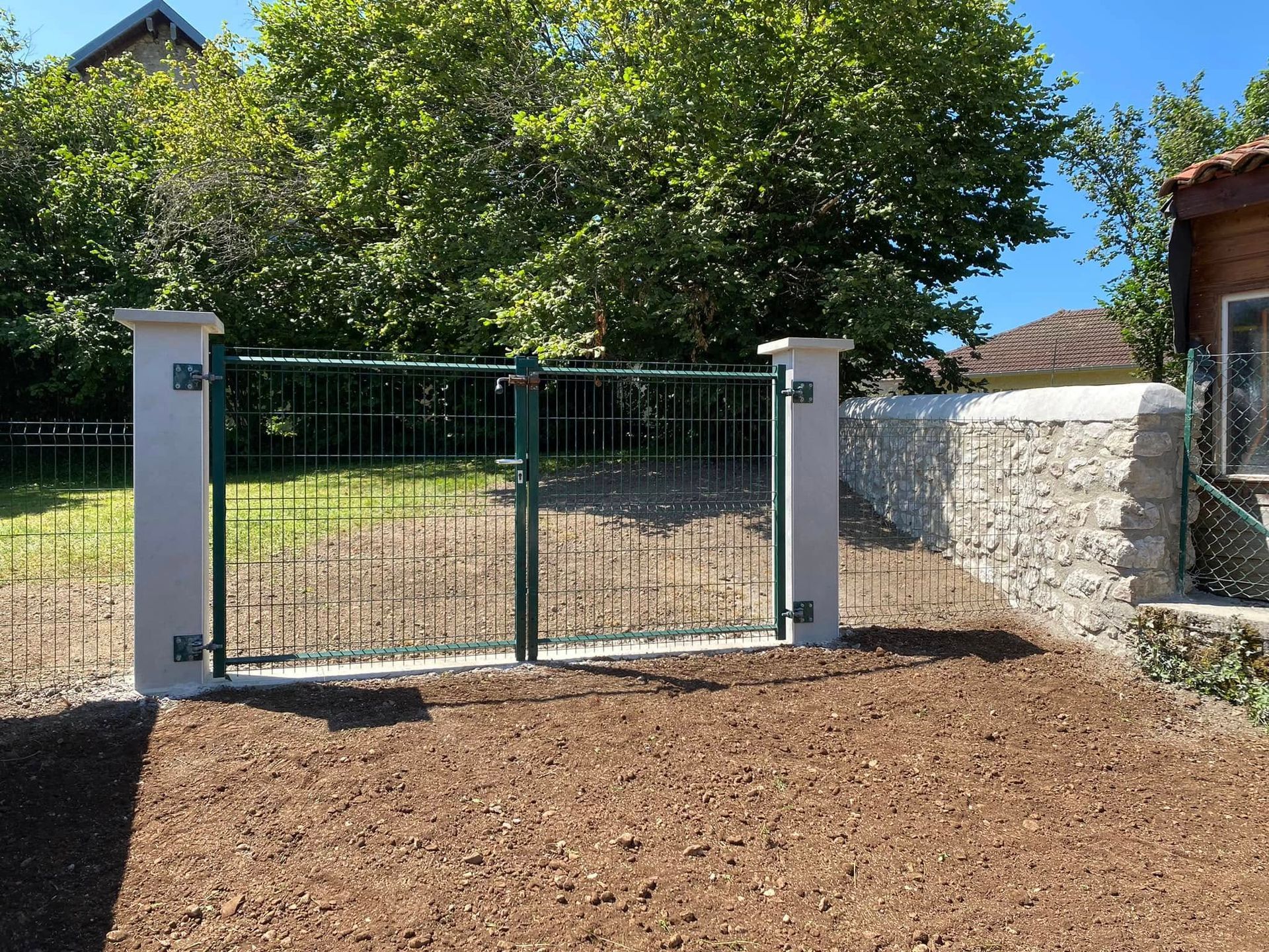 Un portail en métal vert entre des piliers blancs marque l'entrée d'une maison, avec des arbres et un mur de pierre à proximité.