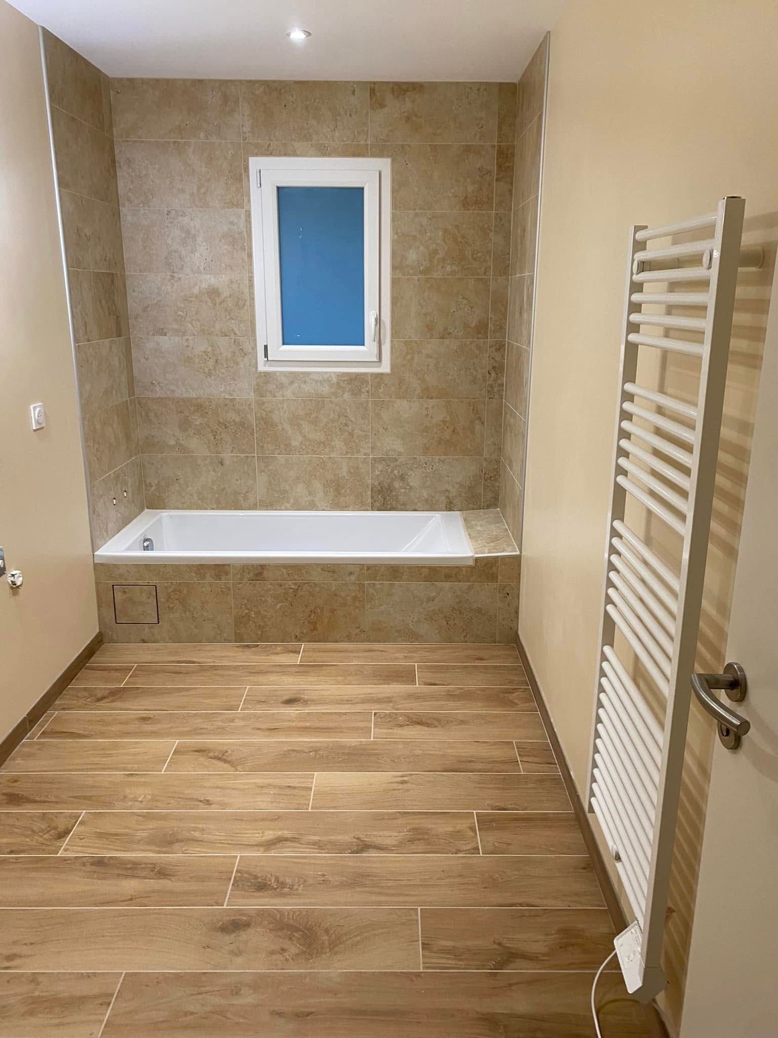 Salle de bains avec sol imitation bois, baignoire encastrée carrelée, petite fenêtre dépolie et sèche-serviettes blanc.