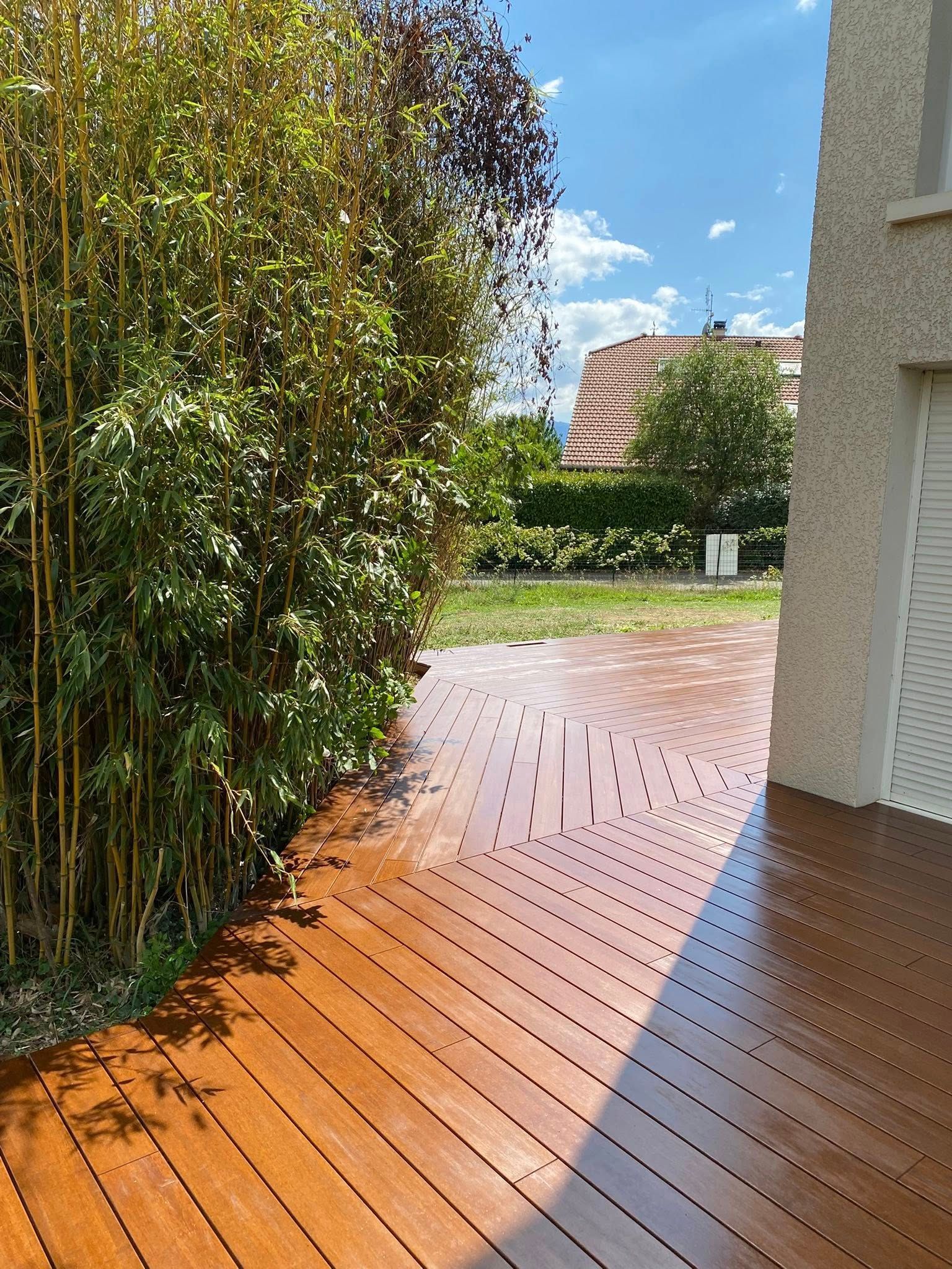 Terrasse en bois jouxtant une haie et une maison, le soleil projetant une ombre incurvée vers une cour gazonnée.