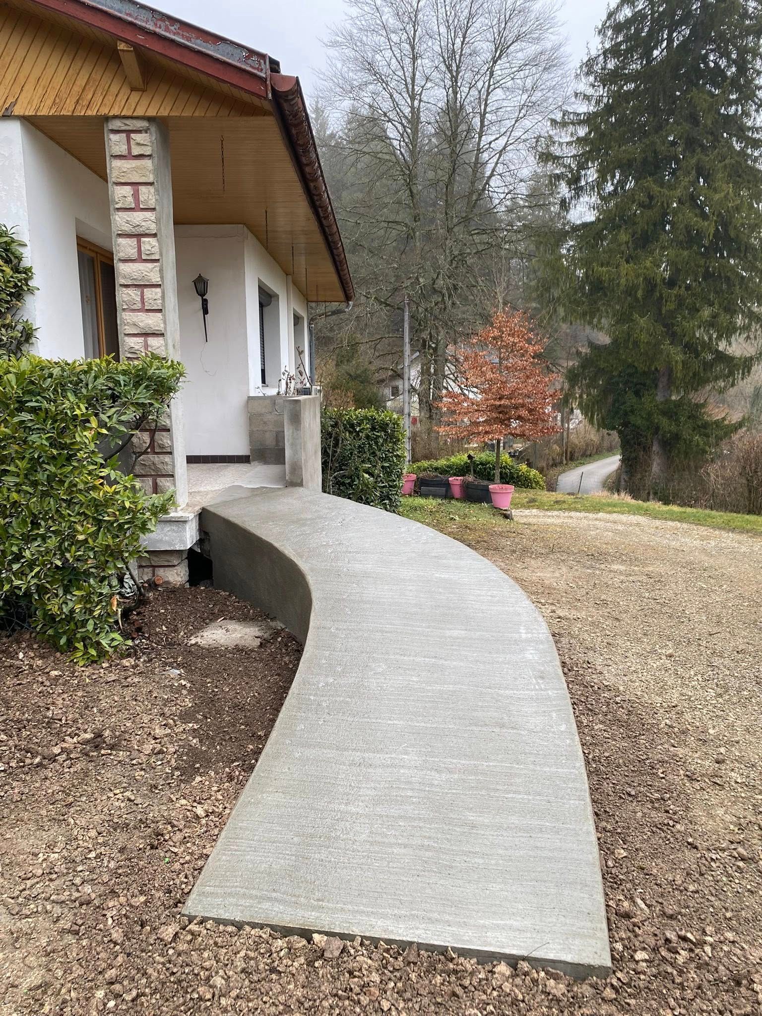 Allée en béton incurvée menant à l'entrée d'une maison, bordée d'arbustes et d'une cour en gravier