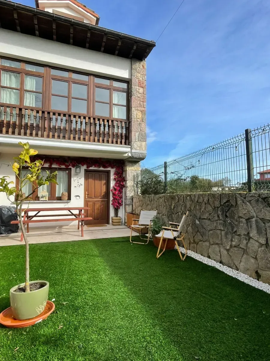 Casa de dos plantas con fachada de piedra, balcón y puerta de madera. Césped con maceta, asientos y muro de piedra.