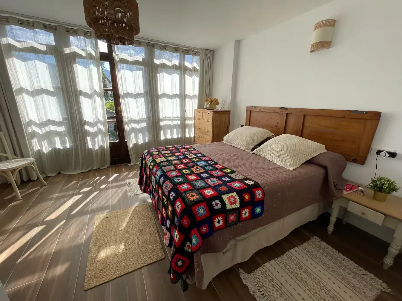 Dormitorio con cabecero de madera, manta de patchwork, cortinas transparentes y luz solar en el suelo.