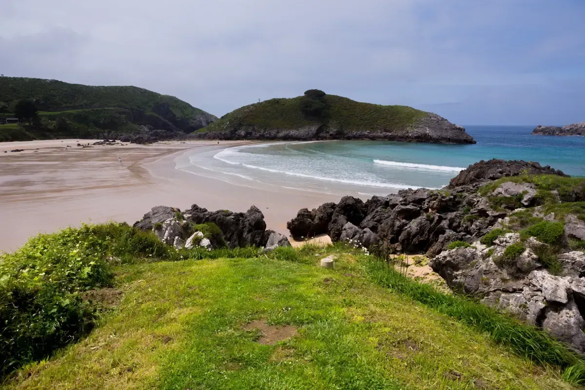 Playa de arena y agua turquesa, con césped en primer plano y colinas verdes en el fondo.