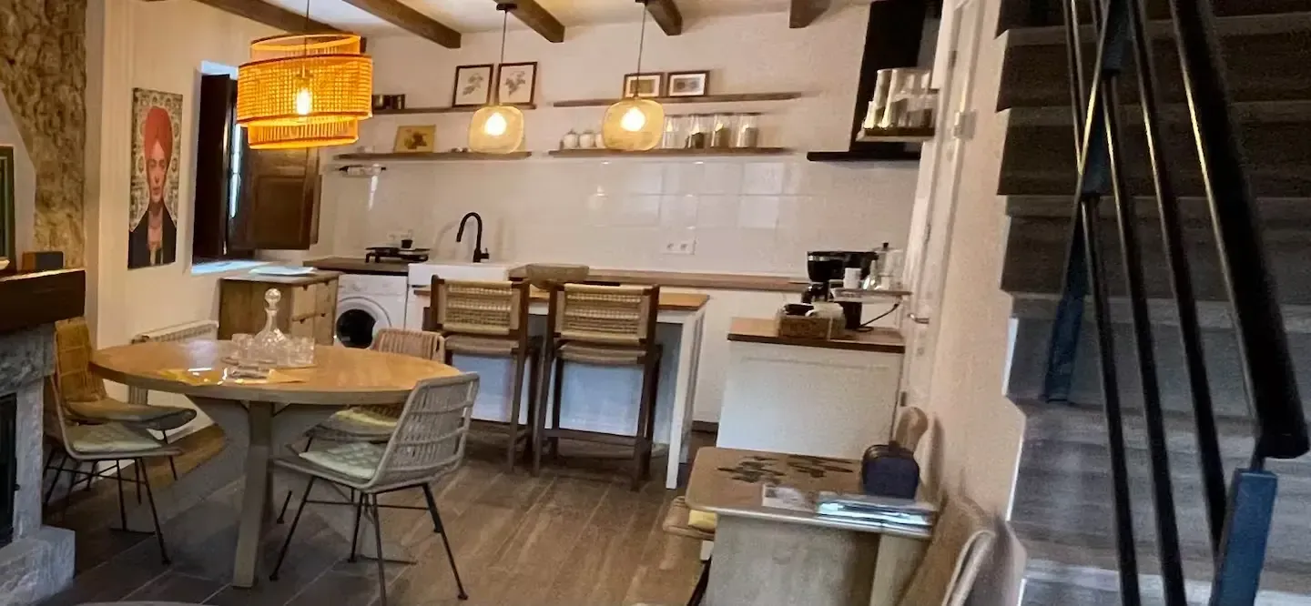 Cocina rústica con mesa de comedor, barra americana y escalera. Iluminación cálida y vigas de madera.