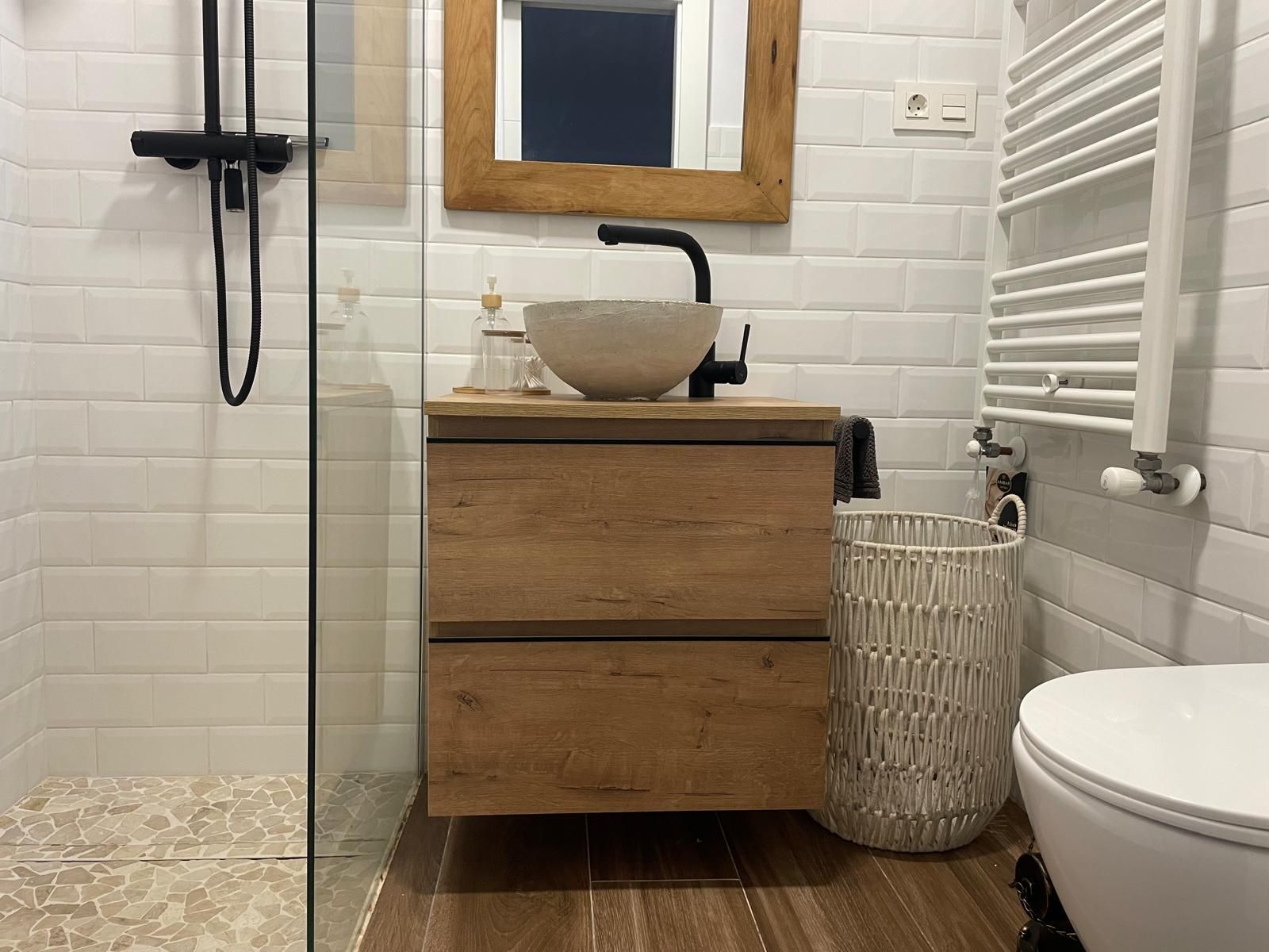 Baño con tocador de madera, lavabo sobre encimera, azulejos tipo metro y puerta de ducha de vidrio.