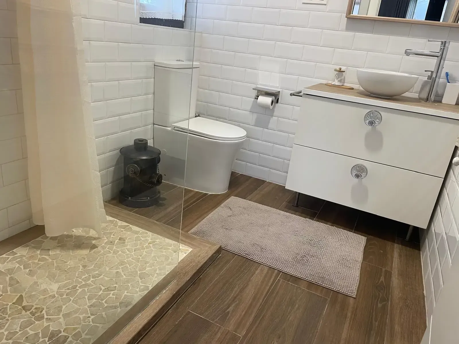 Baño con paredes de azulejos blancos, inodoro, tocador, ducha y una pequeña alfombra sobre el suelo con aspecto de madera.