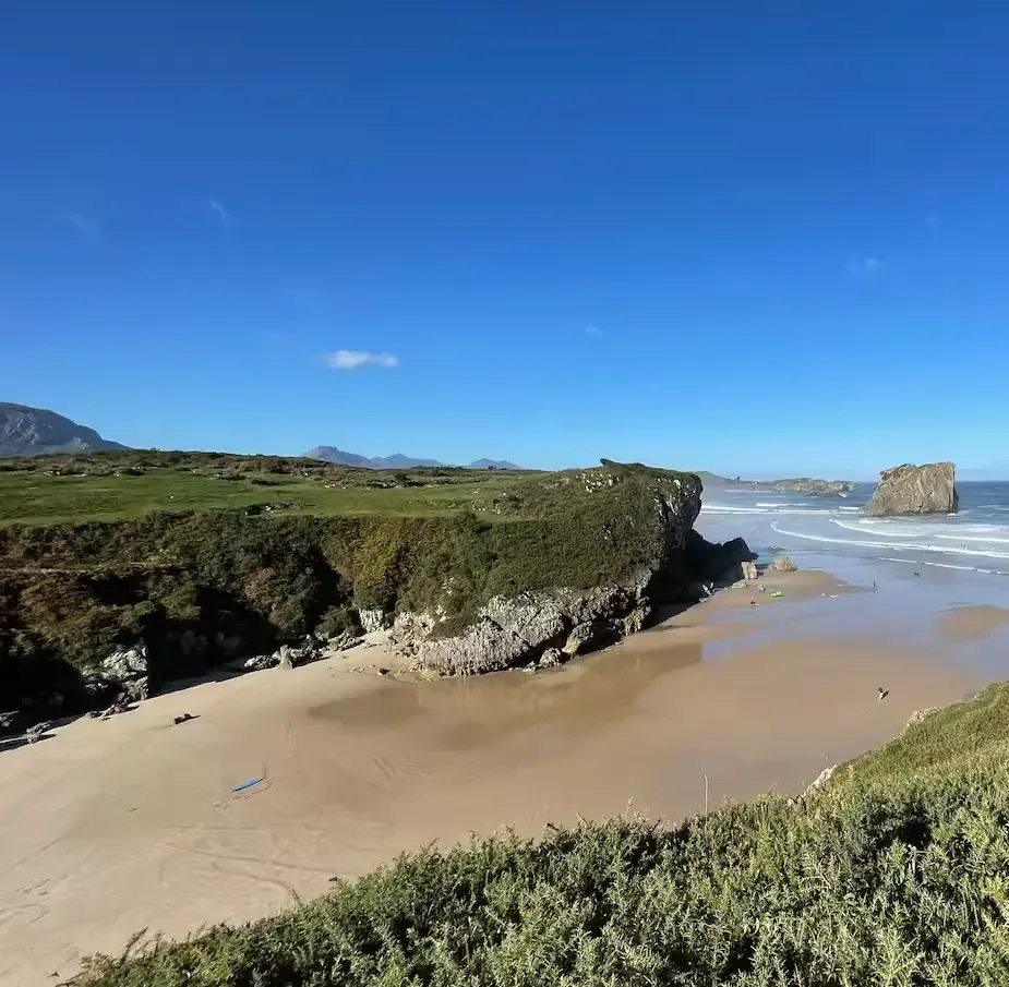 Playa de arena con olas del océano, acantilados cubiertos de hierba y un cielo azul brillante.
