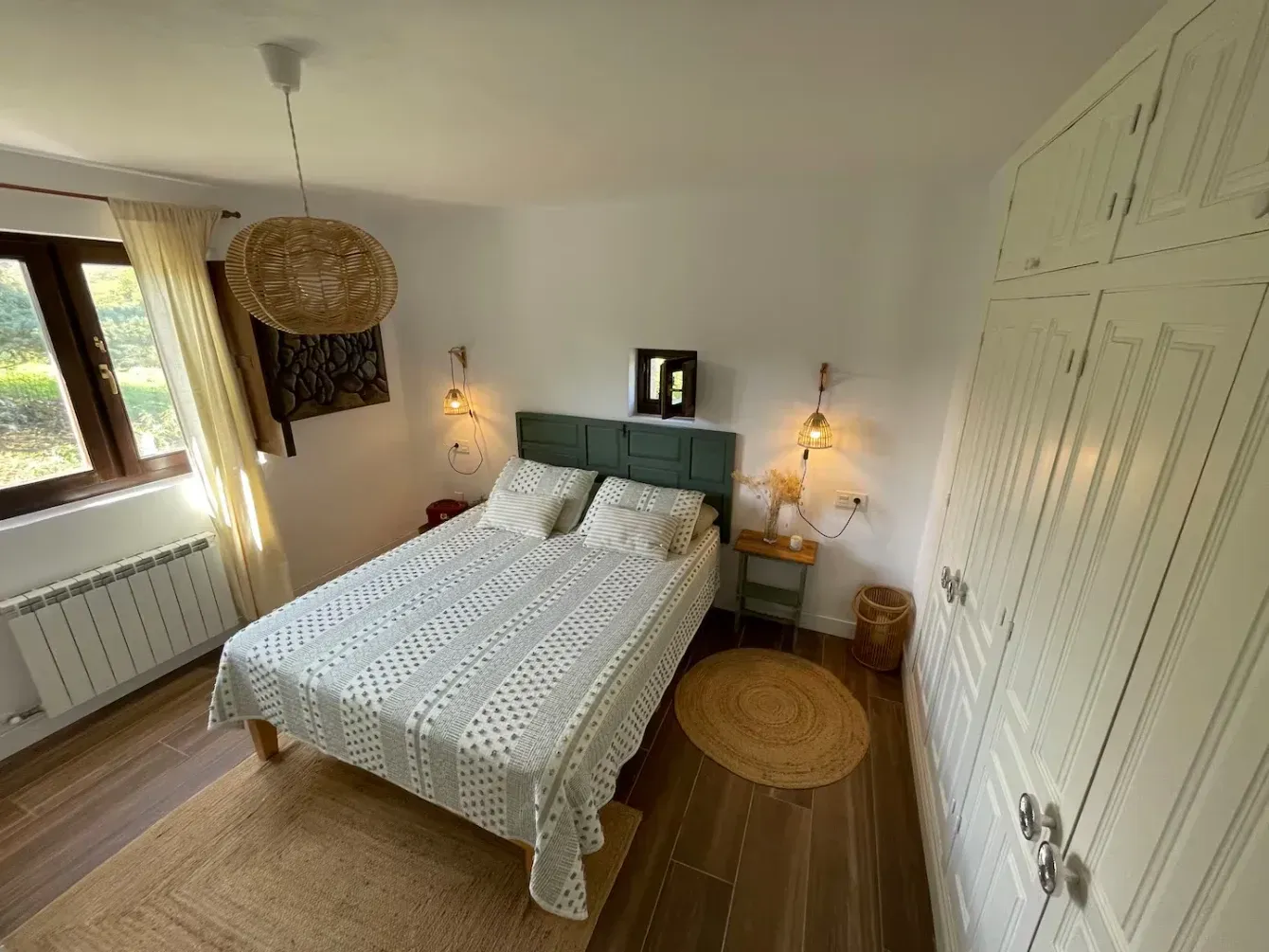 Dormitorio con cama doble, armario blanco y alfombra tejida. Luz natural que entra por la ventana.