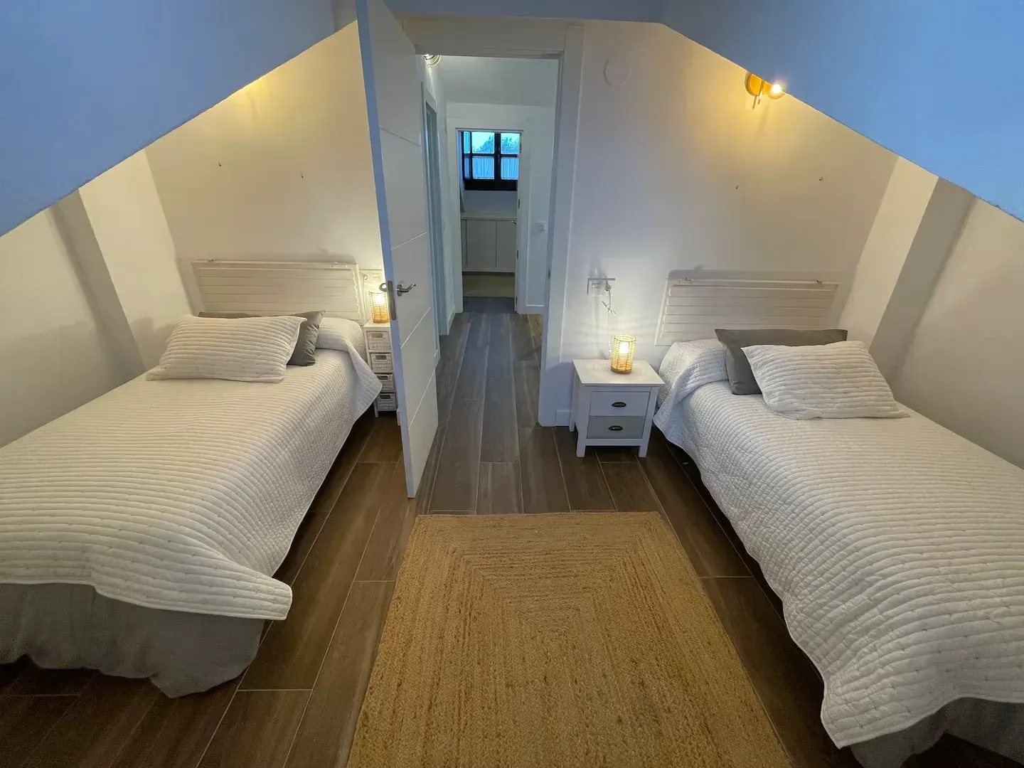 Dos camas individuales en una habitación luminosa con alfombra y puerta abierta que conduce a una cocina.