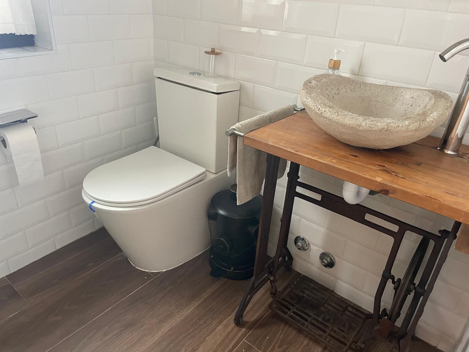 Un pequeño baño con paredes de azulejos blancos, un inodoro y un lavabo sobre una mesa de madera y metal.