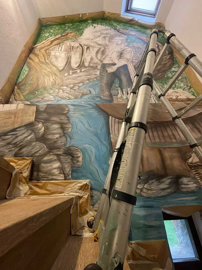 Escalera interior con una pared decorada con un mural, barandillas metálicas y una escena de un arroyo rocoso bajo un tragaluz