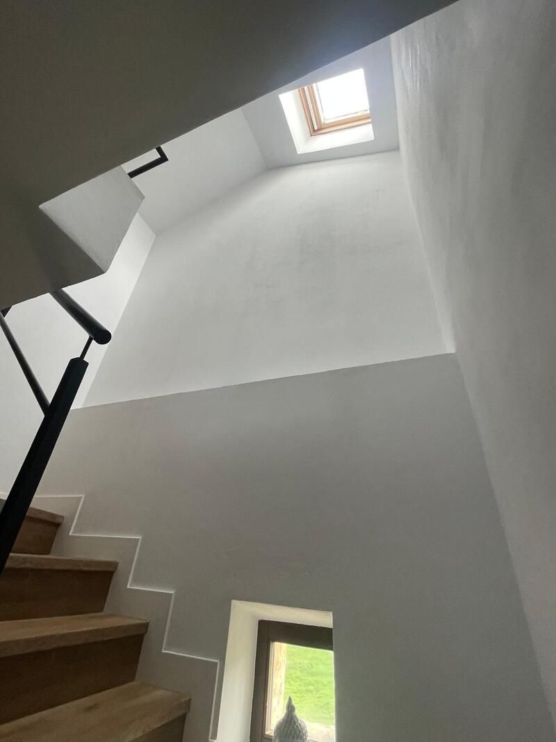 Escalera con paredes blancas, peldaños de madera, barandilla negra y tragaluz en la parte superior.