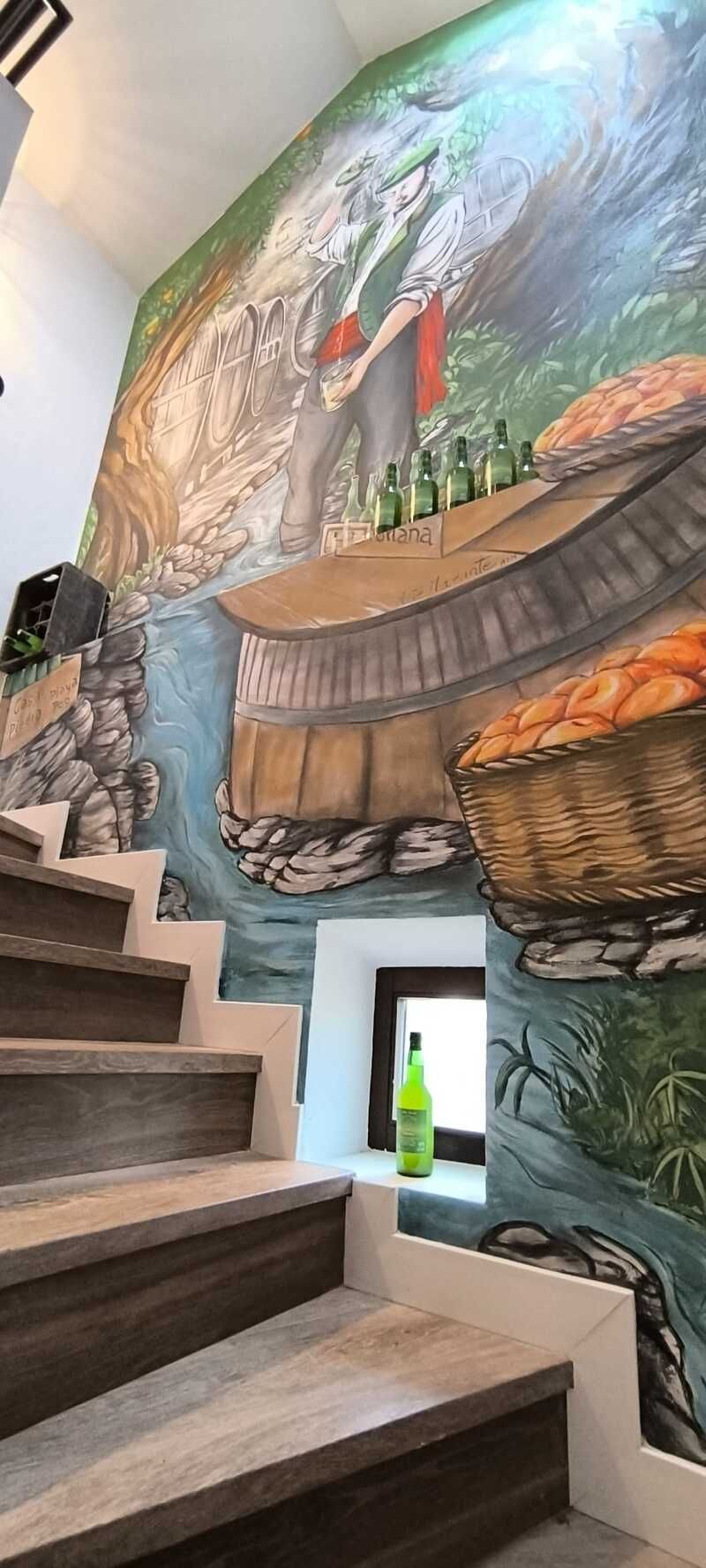 Escalera junto a un colorido mural de un pueblo y montañas, con una pequeña ventana en la que se ve una botella verde.