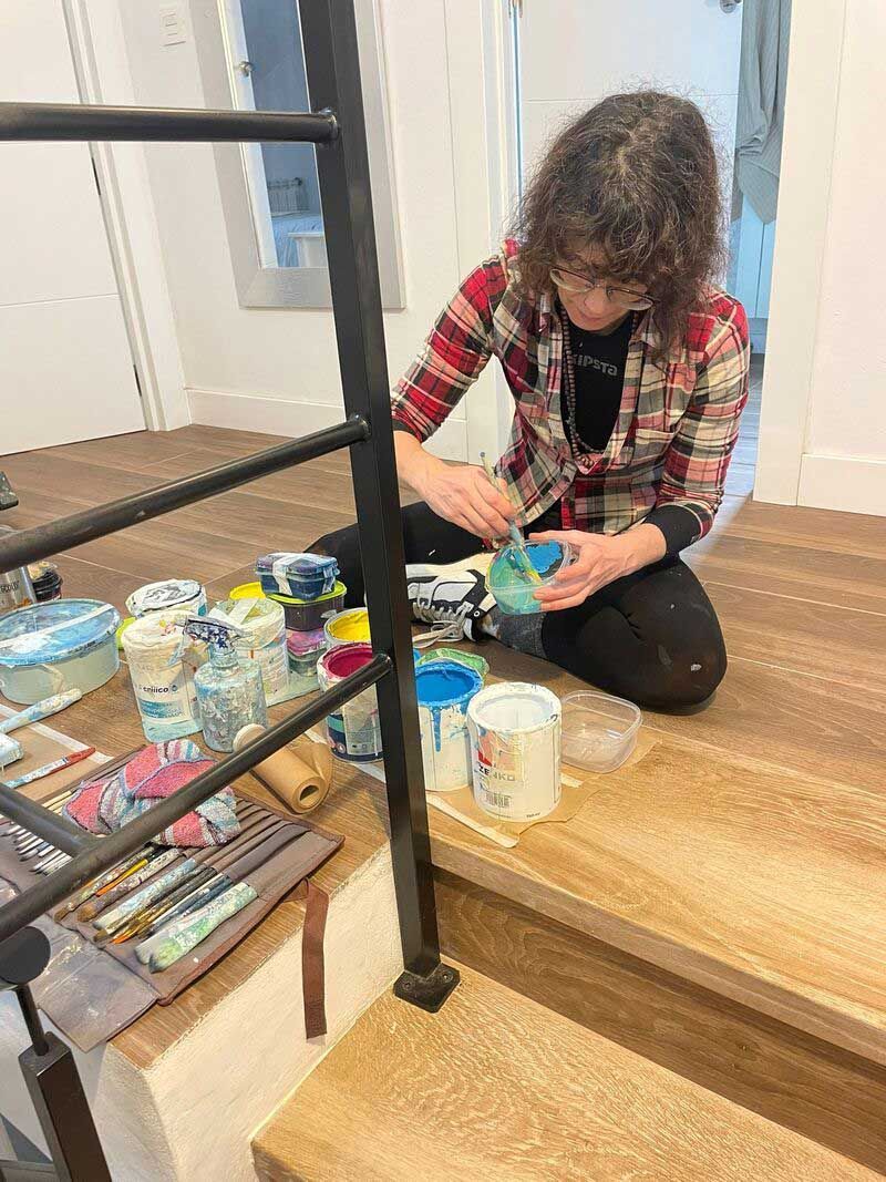Persona arrodillada en un escalón alfombrado, pintando un recipiente pequeño junto a materiales de arte y una mesa.