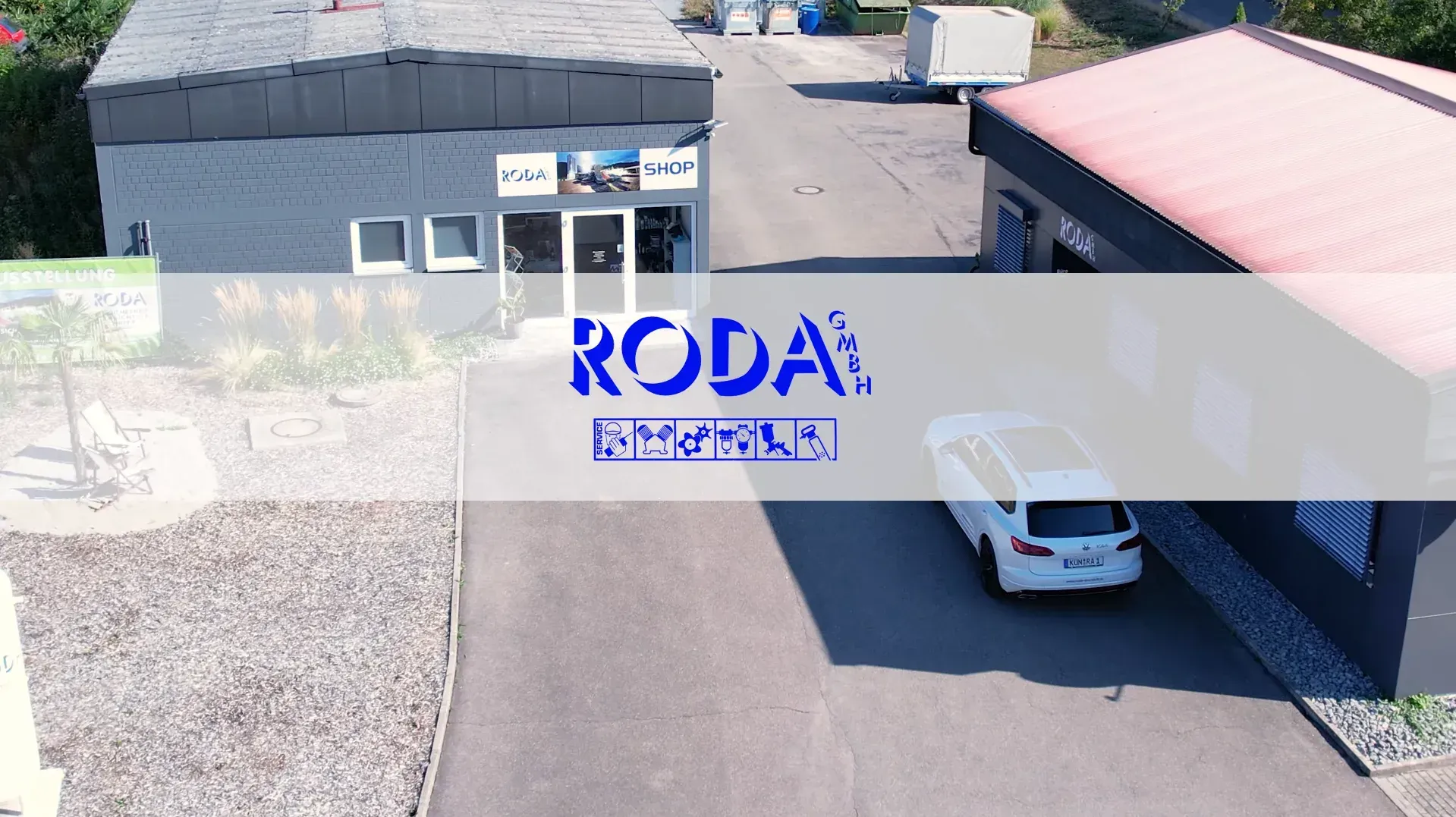 Luftaufnahme eines Gebäudeeingangs mit einem weißen Auto, das unter einem Vordach geparkt ist, und einem großen RODA-Logo darüber.