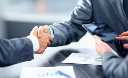zwei Personen im Anzug schütteln sich die Hände, dritte Person tippt am Handy