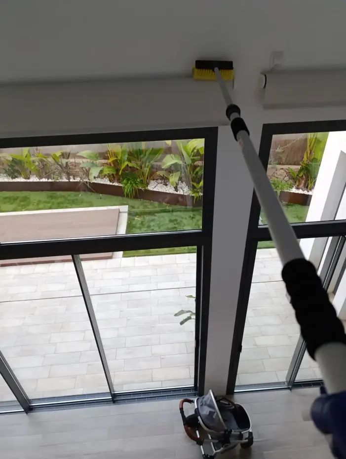 Persona que utiliza un plumero telescópico para limpiar un techo blanco en el interior, cerca de ventanas de vidrio que dan a un patio.