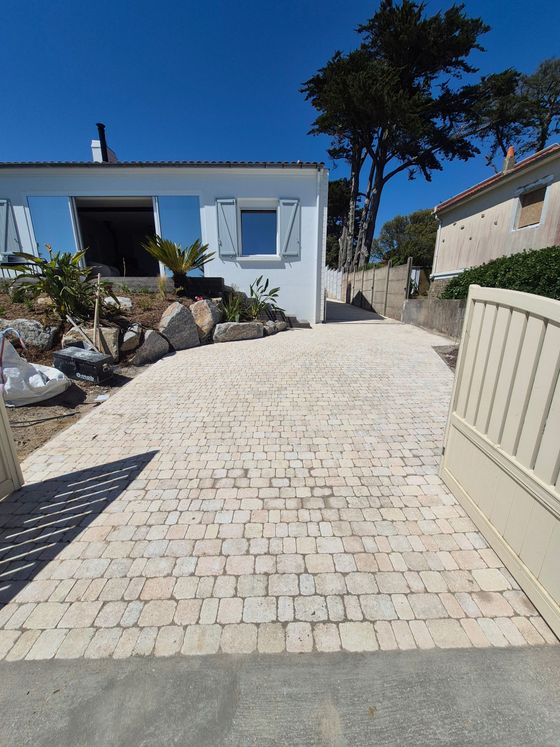 Maison moderne en pierre avec garage triple, longue allée et arbres taillés sous un soleil radieux.