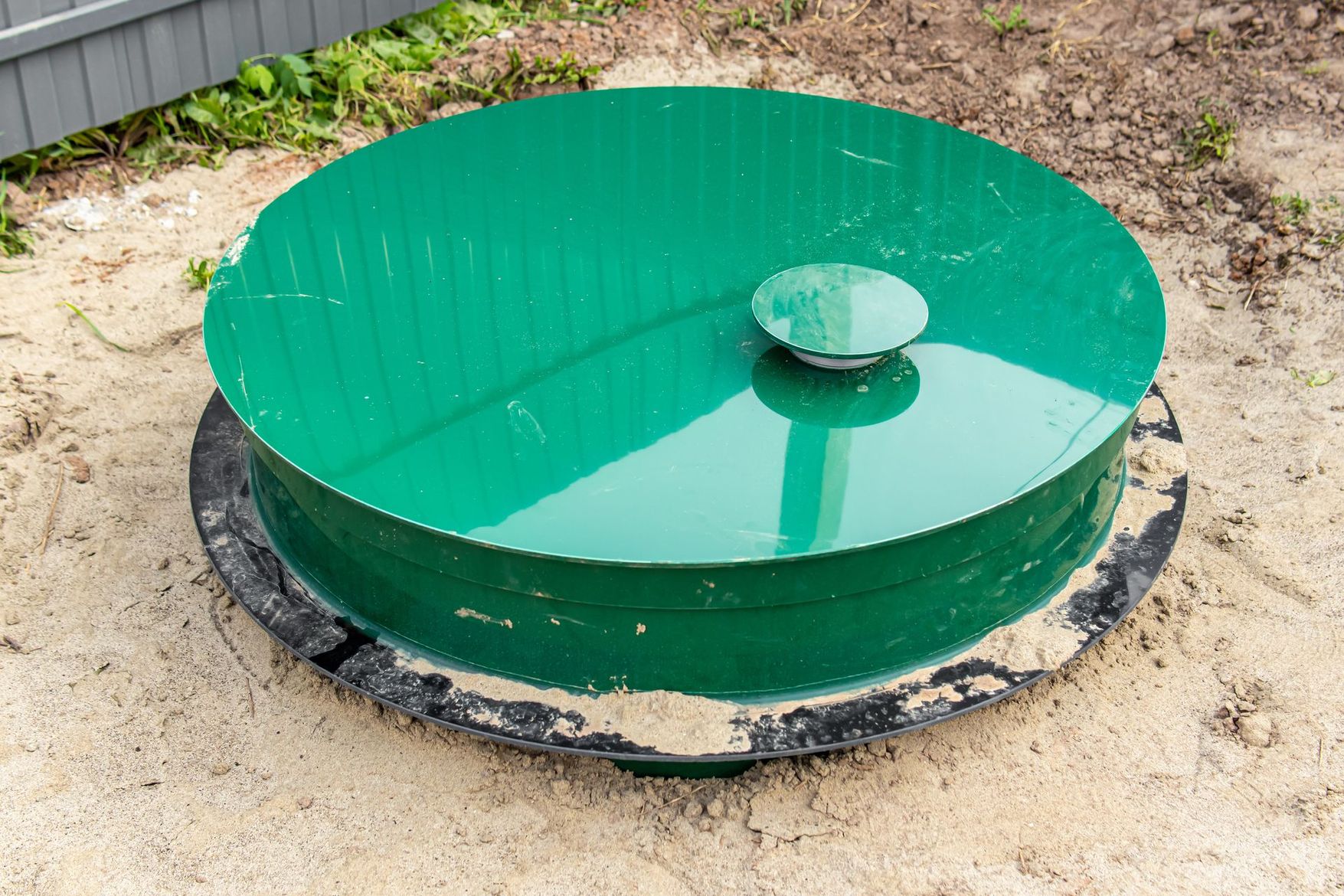 Couvercle utilitaire circulaire en métal vert, dans la saleté, avec un capuchon central surélevé.
