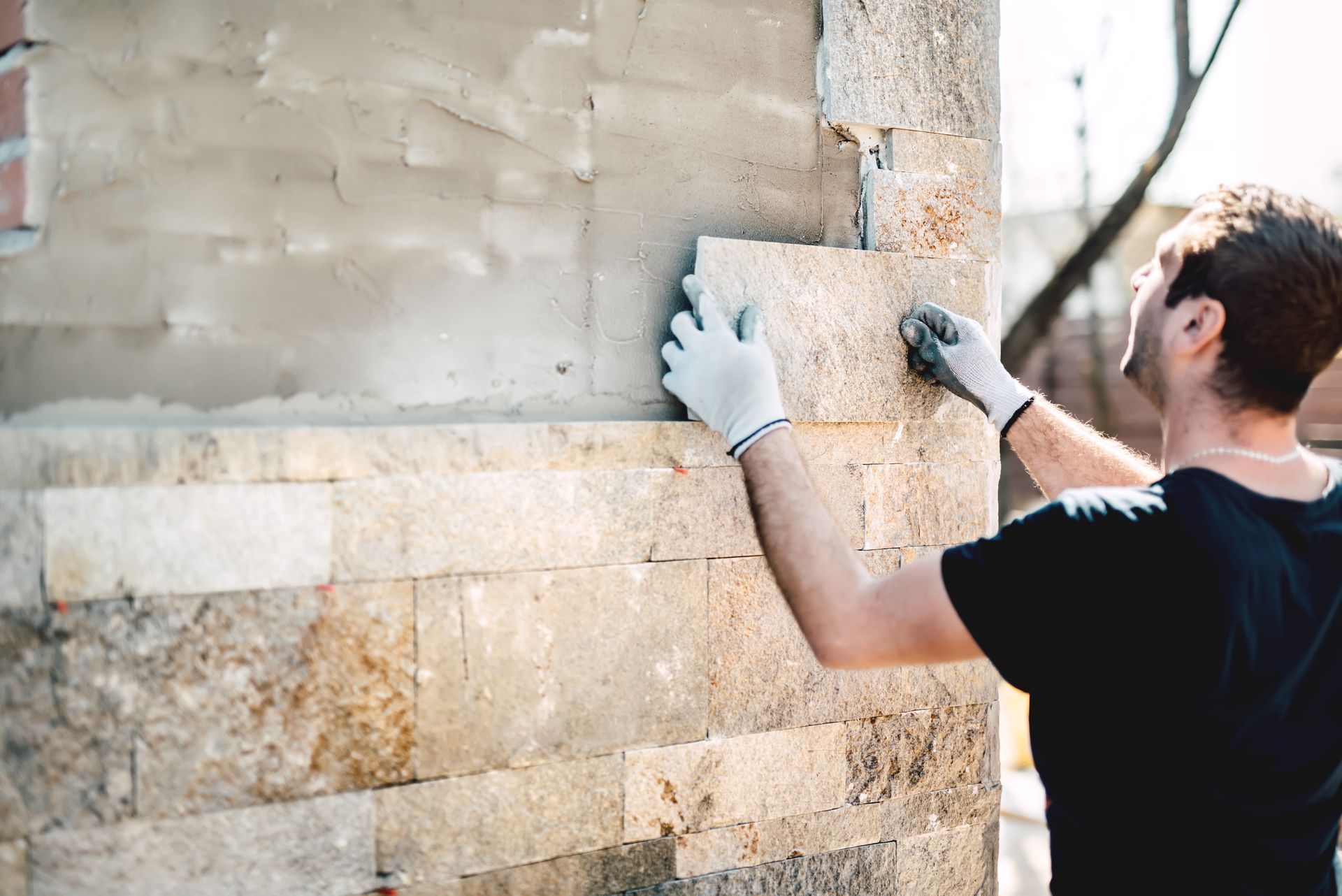 Une personne installe un mur en parement de pierre à l'extérieur, à l'aide de gants et d'une truelle.