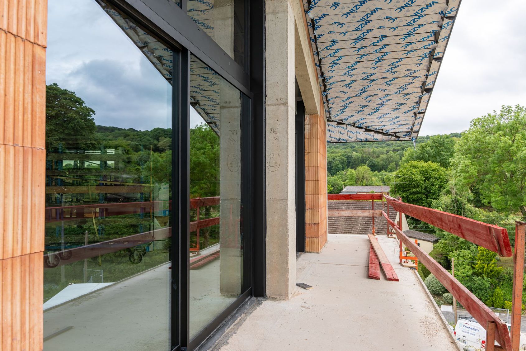 Balcon avec portes vitrées, sol en béton et balustrades rouges donnant sur des arbres verdoyants