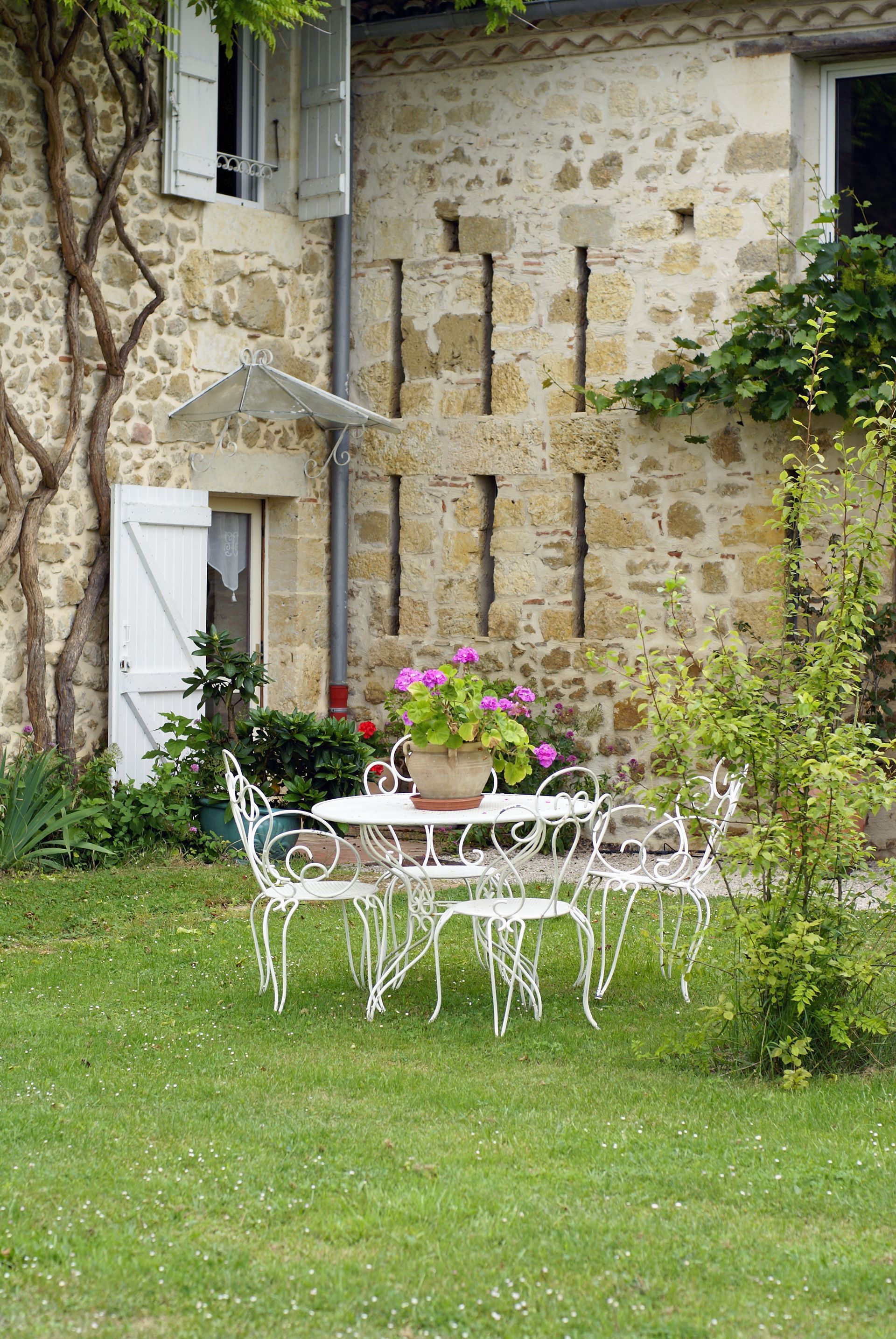Table et chaises en fer forgé blanc avec des fleurs roses dans un jardin jouxtant une maison en pierre.