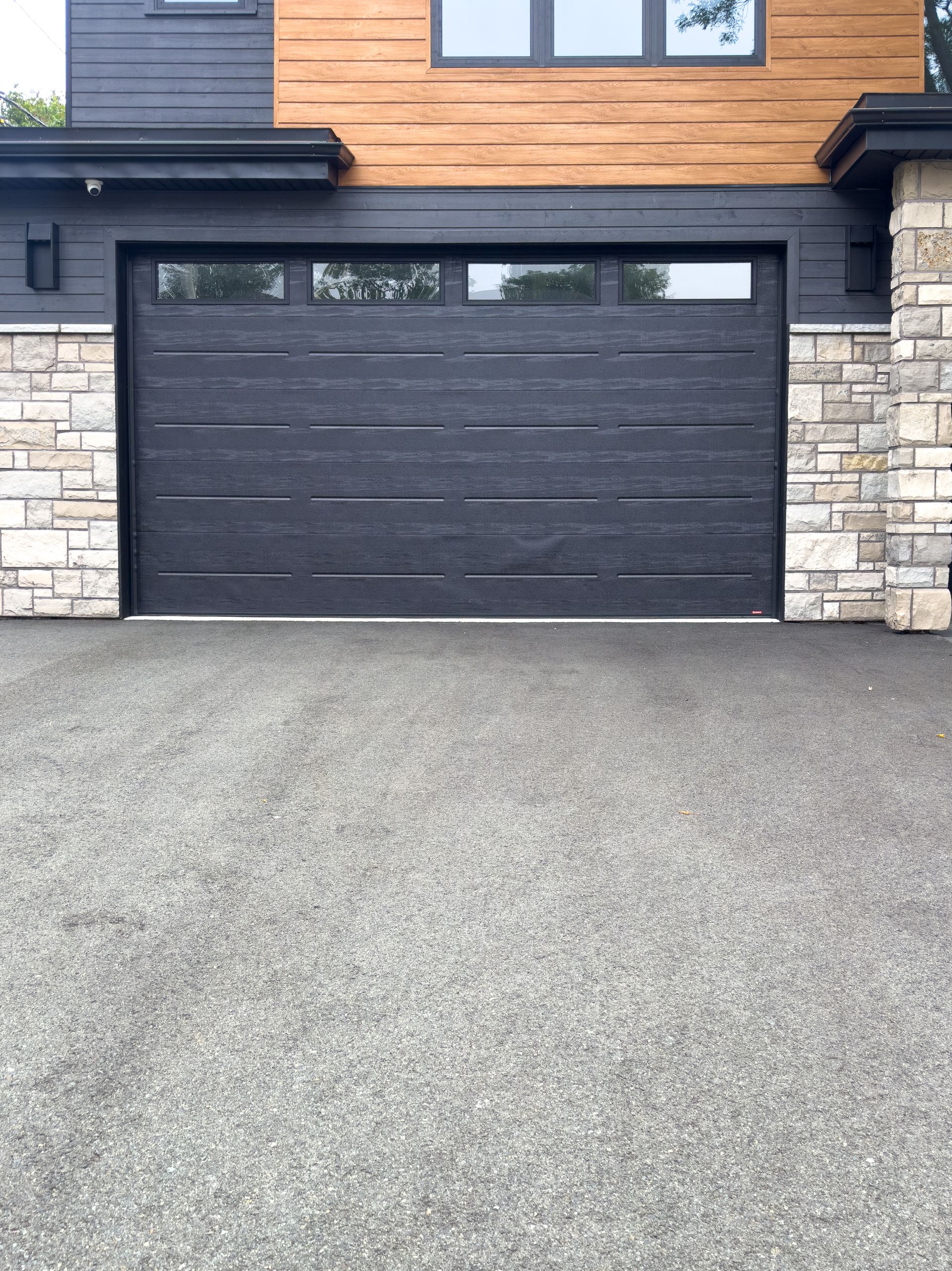 Extérieur de maison moderne avec une porte de garage noire, des éléments en pierre et une allée pavée.