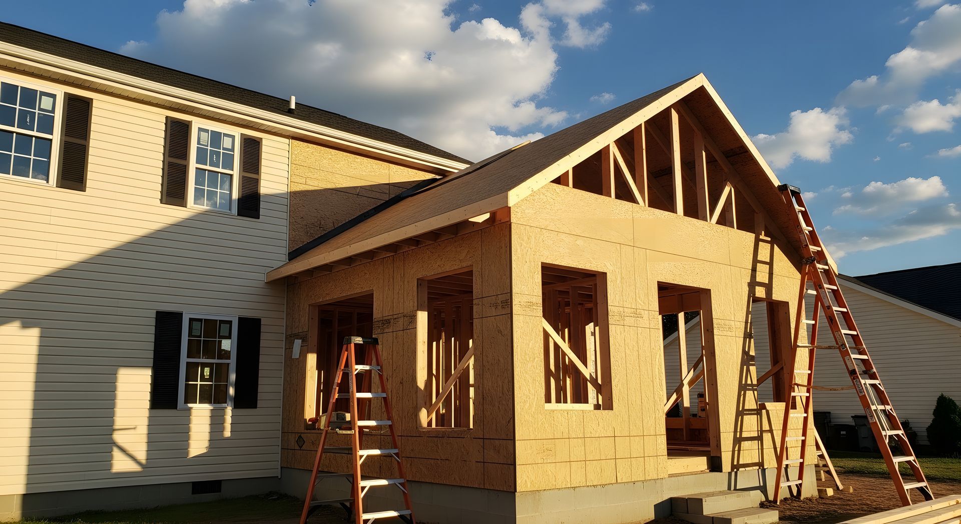 Une extension de maison est en construction à côté d'une maison couleur crème, avec une charpente apparente et des échelles.