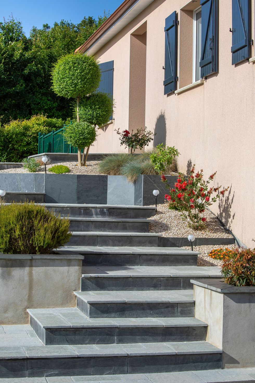 Chemin de pierres en escalier longeant une maison beige aux volets bleus et aux fleurs en pot, montant vers la colline