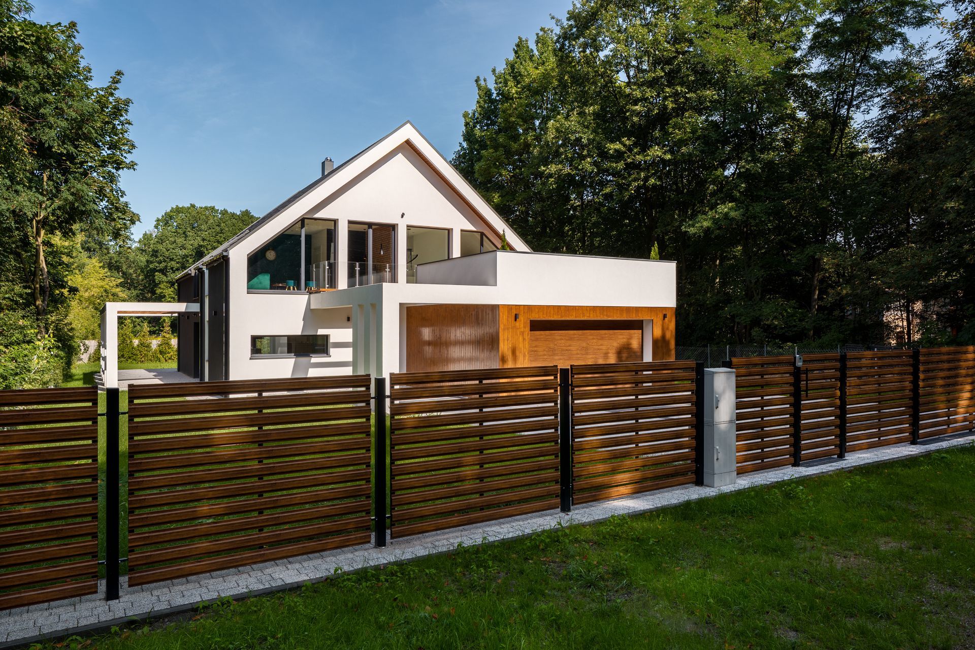 Maison moderne derrière une clôture en bois sombre, avec des arbres et une pelouse verte.