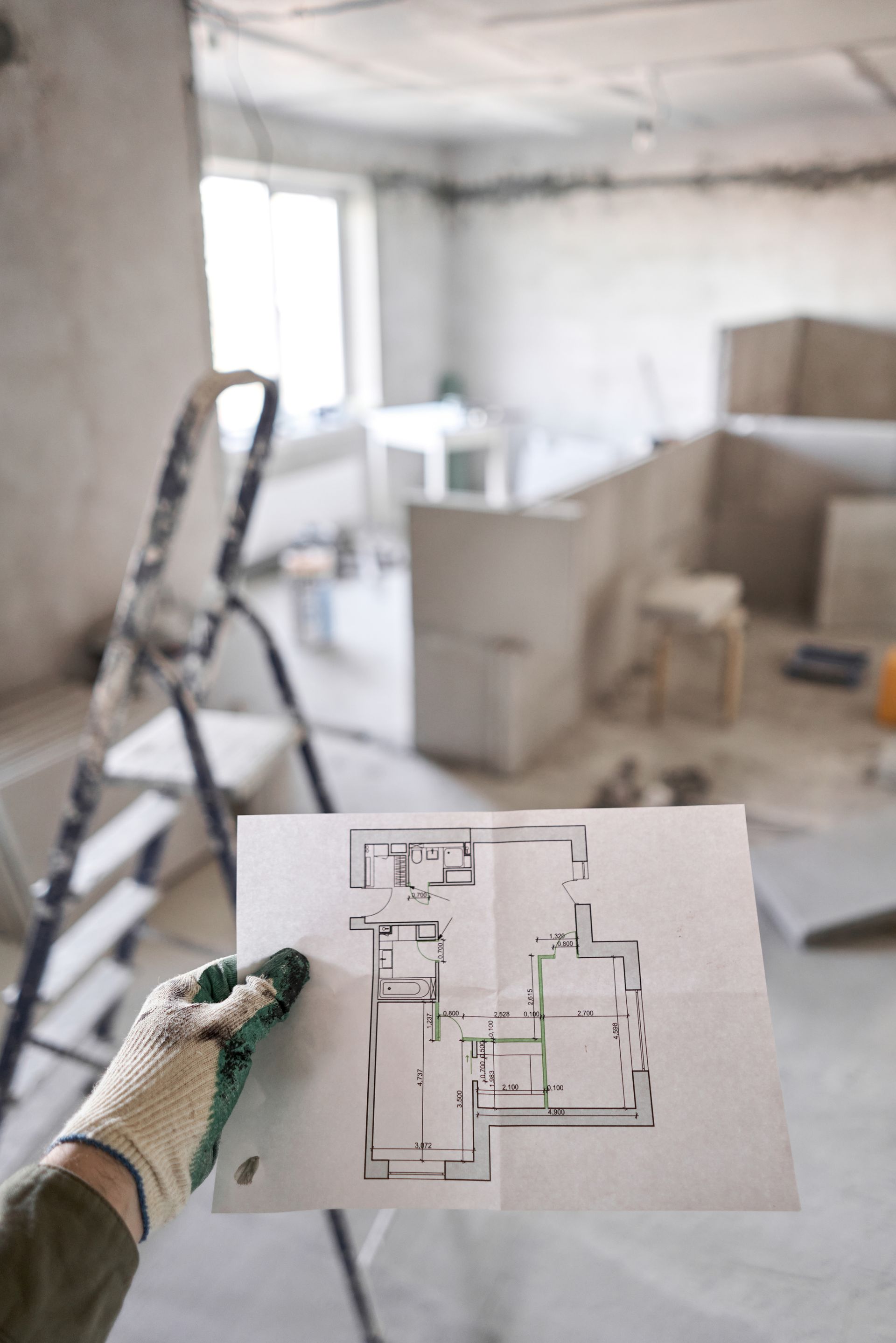 Une main gantée tenant un plan d'étage dans une pièce poussiéreuse et inachevée, avec des outils.