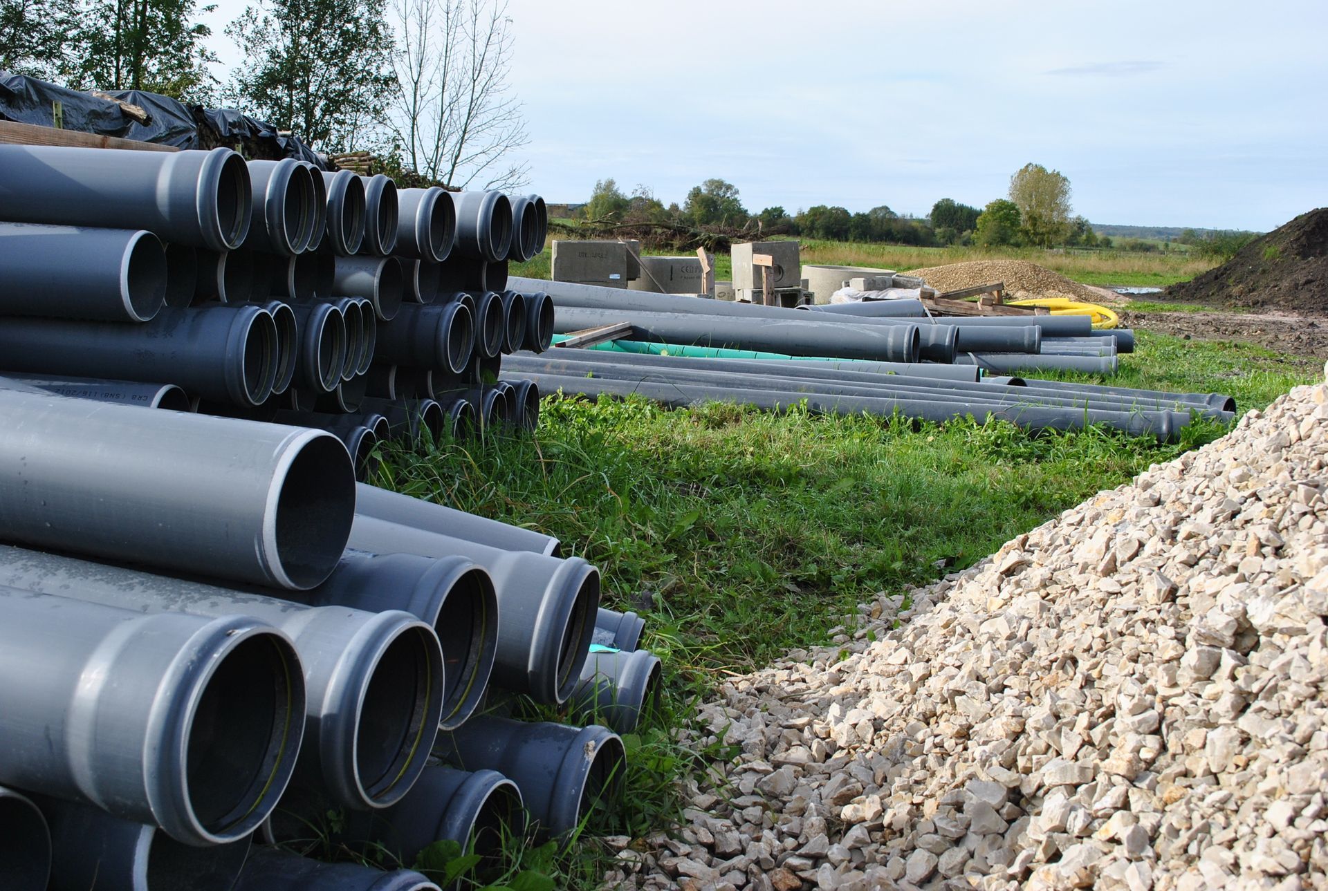 Des piles de gros tuyaux de drainage gris jouxtent un tas de gravier sur un chantier de construction en plein air.
