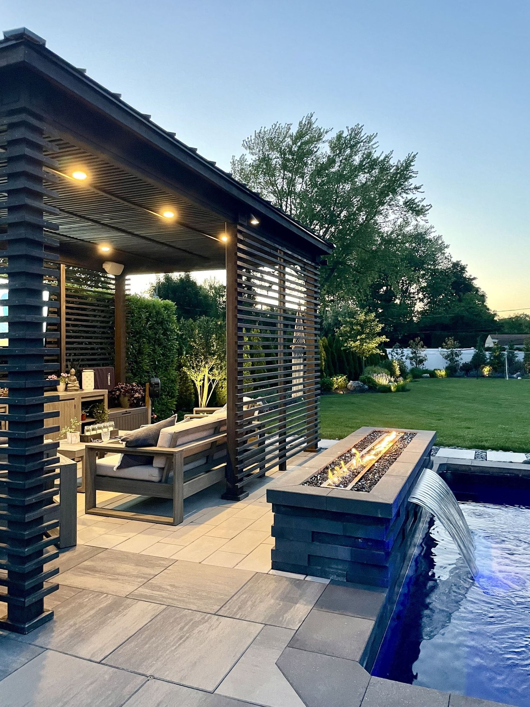 Pergola en bois au bord d'une piscine éclairée de bleu au crépuscule, avec une longue table.